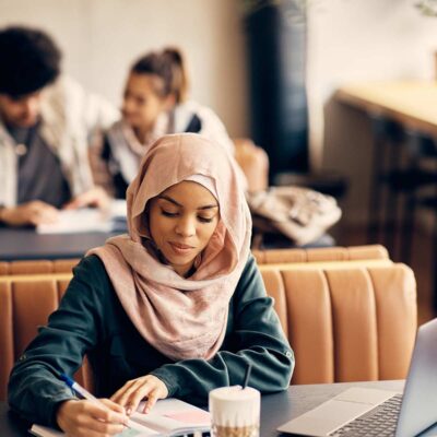 Middle Eastern woman writing notes while studying on laptop at campus cafeteria